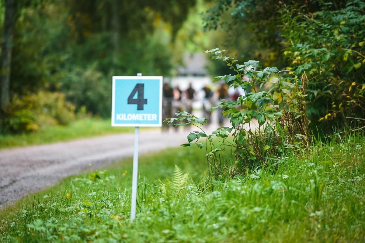 Schild „4 Kilometer“ am Waldrand, Fahrergruppe unscharf im Hintergrund