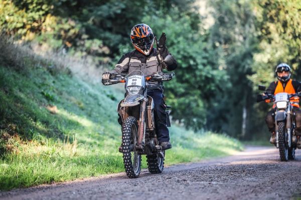 Schlussfahrer auf einem Motorrad fährt auf einem Schotterweg und winkt.