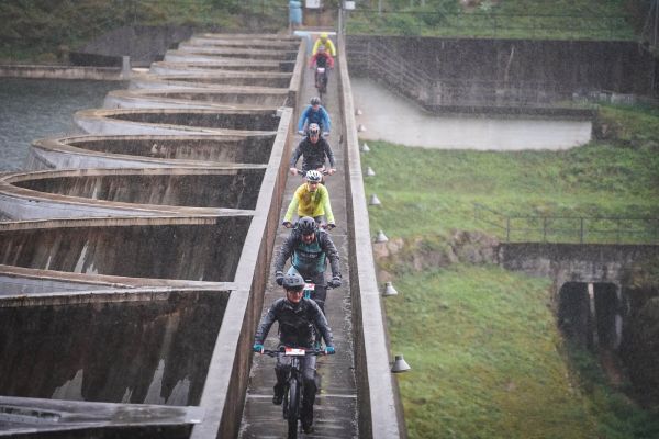 Mountainbiker fahren in einer Reihe über eine schmale Brücke bei starkem Regen