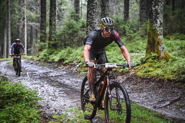 Verschlammter Mountainbiker auf nassem Waldweg, ein weiterer Fahrer folgt